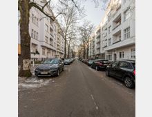 Straßenansicht mit gepflastertem Asphaltweg, gesäumt von parkenden Autos und mehrstöckigen Wohngebäuden auf beiden Seiten. Die Gebäude sind hell und haben Balkone, kahle Bäume ragen über die Straße.