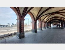 Arkadengang der Oberbaumbrücke in Berlin mit rot-schwarz gemusterten Backsteinbögen und spitz zulaufenden Gewölben. Im Hintergrund sind moderne Gebäude und die Spree zu sehen.