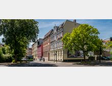 Straßenszene mit historischen Wohngebäuden, darunter ein rotes Backsteinhaus mit weißen Fensterrahmen und Zierelementen, flankiert von grünen Bäumen. Im Vordergrund sind ein Fußgängerüberweg, Fahrräder und ein Bus an einer Kreuzung sichtbar.