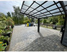 Überdachter Stellplatz mit gepflasterter Fläche, flankiert von Blumenkübeln auf der linken Seite und einer Müllstation mit farbigen Tonnen im Hintergrund. Im Hintergrund sind ein Zaun, Bäume und eine Wohnstraße sichtbar.