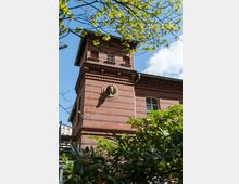 Backsteinbau mit Turm und verzierten Fenstern, umgeben von grünem Laub, unter einem blauen Himmel mit wenigen Wolken. Ein Relief rund um ein kreisförmiges Element ist an der Fassade sichtbar.