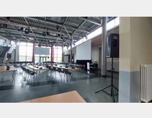 Großer Tagungsraum mit einer modernen Einrichtung, bestehend aus gestellten Tischen und Stühlen, mehreren Mikrofonen, zwei großen Projektionsflächen und Lautsprechern. Der Raum hat eine hohe Decke mit sichtbaren Stahlträgern und große Fensterfronten, die viel Tageslicht hereinlassen.