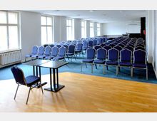 Ein moderner Konferenzraum mit mehreren Reihen von blauen Stühlen auf blauem Teppichboden. Im Vordergrund steht ein einzelner Tisch mit einem Stuhl auf einem Parkettbereich, dahinter große Fenster, die Tageslicht hereinlassen.