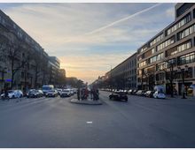 Straßenszene in einer Stadt bei Sonnenuntergang mit symmetrisch verlaufenden Fahrbahnen, flankiert von mehrstöckigen Gebäuden auf beiden Seiten. Autos stehen im Verkehr, und Fußgänger überqueren die Straße, mit kahlen Bäumen entlang der Gehwege.