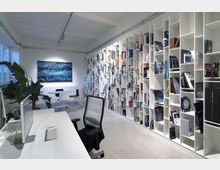 Modernes Büro oder Arbeitsraum mit einer Rückwand aus hohen, weißen Bücherregalen, die mit Büchern und Zeitschriften gefüllt sind. Im Vordergrund steht ein weißer Schreibtisch mit einem Computer, Stuhl und im Hintergrund ein kleiner Besprechungsbereich mit Tisch und Stühlen sowie einer Zimmerpflanze.