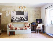 Ein schlichtes Zimmer mit abgenutzter Wand, einem weißen Tagesbett mit rot gemusterten Kissen und Decken, sowie einem kleinen Holztisch mit einem Laptop, Monitor und weißem Stuhl. Auf dem Bett hängt ein großes schwarz-weiß-Fotografiedruck, und am rechten Rand befindet sich ein Fenster mit weißen Vorhängen.