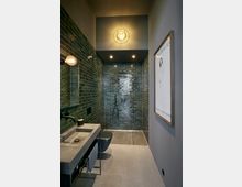 Minimalistisch gestaltetes Badezimmer mit einer bodentiefen Dusche, deren Wände mit glänzenden dunkelgrünen Fliesen verkleidet sind. Der Raum enthält ein Betonwaschbecken, einen schwarzen Wasserhahn, graue Bodenfliesen und eine Wandleuchte an der Decke.