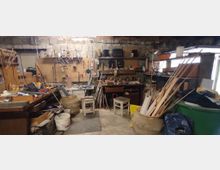 Werkstatt mit chaotisch angeordneten Arbeitsmaterialien und Werkzeugen. Holzleisten, Werkbänke, Regale mit verschiedenen Utensilien sowie ein grober Betonboden und eine unverputzte Steinmauer prägen den Raum.