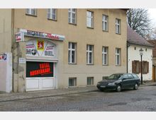 Straßenansicht eines zweistöckigen Gebäudes mit gelber Fassade und mehreren Fenstern. Im Erdgeschoss befindet sich ein Geschäft mit der Aufschrift "Möbel Markt" und einem Schild, das auf einen "Total Ausverkauf" hinweist; davor steht ein geparktes Auto auf einer gepflasterten Straße.