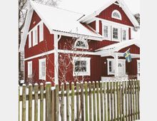 Rotes, zweistöckiges Holzhaus im schwedischen Stil mit weißer Fenster- und Türumrandung, umgeben von einer dünnen Schneedecke. Im Vordergrund steht ein Holzzaun, und an der Veranda ist eine schwedische Flagge befestigt.