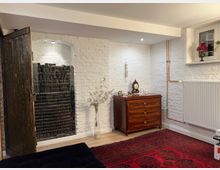 Ein gemütlicher Innenraum mit weiß gestrichenen Backsteinwänden, einer kleinen Holzkommode mit antiker Uhr und Dekoration darauf sowie einem roten Teppich mit geometrischen Mustern. Rechts an der Wand befindet sich ein Heizkörper, links eine geöffnete Tür aus dunklem Holz, die ein Regal mit Büchern und Gegenständen im Hintergrund freigibt.