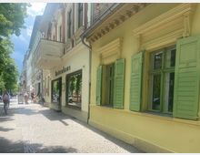 Straßenszene mit mehrstöckigen Altbauten, darunter ein Geschäft mit dem Namen „Bettehaus“. Die Fassade eines Gebäudes rechts ist gelb gestrichen und mit grünen Fensterläden versehen. Links verlaufen Bäume entlang des Gehwegs, auf dem Fußgänger und ein Radfahrer zu sehen sind.