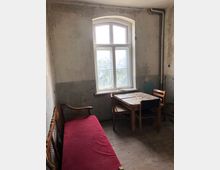 Ein einfacher Raum mit abgenutzten Wänden und einem großen, bogenförmigen Fenster in der Mitte. Im Vordergrund stehen ein rotes Sofa und ein Holztisch mit zwei Holzstühlen, der Boden ist aus Holzplanken.