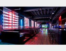 Nachtclub-Innenraum mit dunklem Holzfußboden, beleuchteten Sitzbereichen und bunten LED-Leuchttafeln an den Wänden. Eine lange Bar mit Barhockern befindet sich rechts, während der Raum von farbigem Licht und moderner Architektur geprägt ist.