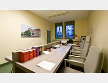 Ein schlicht eingerichtetes Büro mit einem langen Holztisch, mehreren gepolsterten Stühlen und juristischen Fachbüchern. Im Hintergrund sind große Fenster mit Blick auf kahle Bäume sowie eine rote Schreibtischlampe auf dem Tisch zu sehen.