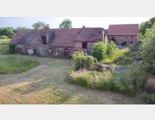 Eine verfallene Scheune aus Backstein mit rotem Ziegeldach, umgeben von überwuchertem Gras und Büschen. Im Hintergrund ist ein weiteres landwirtschaftliches Gebäude zu sehen, ebenfalls mit rotem Ziegeldach.