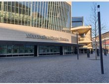 Außenansicht der Mercedes-Benz Arena mit moderner Glas- und Steinarchitektur; im Vordergrund ein gepflasterter Platz mit Laternen und einem kahlen Baum.