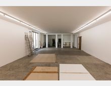 Großer leerer Innenraum mit weiß gestrichenen Wänden, Betonboden und einer langen Reihe Deckenleuchten. An den Seiten stehen Leitern und Werkzeuge; im Hintergrund befinden sich offene Bereiche mit Tischen und Regalen sowie eine Glasfront zur linken Seite.