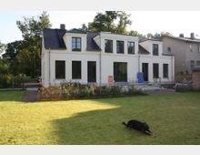 Zweistöckiges, weißes Wohnhaus mit schwarzen Fensterrahmen und einem dunklen Schieferdach, umgeben von einem großen Garten mit einer Rasenfläche und Blumenbeeten. Im Vordergrund liegt ein Hund auf der Wiese, während im Hintergrund Bäume und Teile benachbarter Gebäude sichtbar sind.