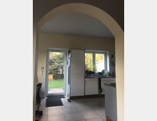 Blick in eine Küche mit hellen Wänden und einem großen runden Durchgangsbogen. Zu sehen sind eine Glastür und ein Fenster mit Blick auf einen Garten, weiße Küchenschränke, eine Arbeitsplatte mit Geschirr sowie eine Fliesenbodenfläche.