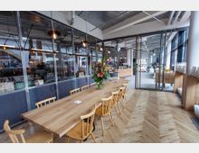 Ein modernes Café oder Restaurant mit großen Glasfenstern, einem langen Holztisch mit Holzstühlen und einem Blumenarrangement in der Mitte. Der Boden ist in Fischgrätmuster verlegt, und die Umgebung zeigt eine offene Gestaltung mit industriellen Elementen wie sichtbaren Rohren an der Decke.