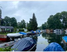 Ein kleiner Hafen mit mehreren an Stegen festgemachten Booten, die größtenteils mit Planen abgedeckt sind. Im Hintergrund sind Bäume, einfache Gebäude und Hausboote am ruhigen Wasser zu sehen.