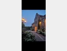 Historisches Gebäude aus Backstein mit mehreren Etagen, von Pflanzen und Blumen umgeben, bei Abenddämmerung beleuchtet. Ein gepflasterter Weg führt entlang blühender Beete, und eine Laterne mit mehreren Leuchten steht links im Bild.