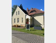 Ein hell verputztes, zweigeschossiges Haus mit rotem Ziegeldach, Dachfenster und Schornsteinen, davor ein kleiner Vorgarten mit Holzbänken, Tisch und zwei Liegestühlen. Im Vordergrund stehen ein Kreidetafel-Aufsteller neben einem Gehweg sowie ein rechteckiger Pflanzkübel am Rasen.