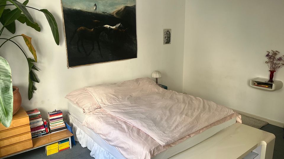 Minimalist bedroom with white walls and a low bed covered in pale pink bedding, with a large dark-toned painting above the headboard. A wooden dresser with stacked books and a large potted plant sits to the left, and a small wall shelf and bedside lamp are on the right.