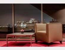 Ein moderner Innenraum mit einem beigen Ledersessel und einem niedrigen rechteckigen Tisch, auf dem eine kleine Vase mit rosa Blumen steht. Im Hintergrund ist durch ein großes Fenster die nächtlich beleuchtete Reichstagskuppel in Berlin sowie das Brandenburger Tor sichtbar.