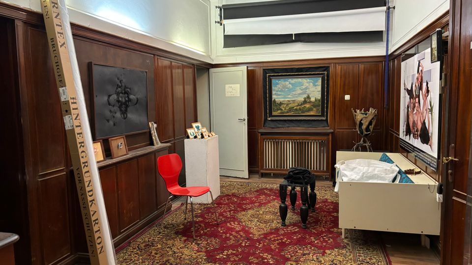 Innenraum eines holzgetäfelten Raums mit hohem weißen Deckenabschluss und großem gemusterten Teppich. An den Wänden hängen gerahmte Kunstwerke; im Raum stehen ein roter Stuhl, mehrere Podeste und Möbelstücke sowie eine Tür und ein Heizkörper an der Stirnwand.