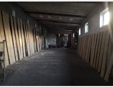 Ein einfacher Innenraum eines langgestreckten Gebäudes mit Betonboden, unverputzten Wänden und einem Holzbalken-Dach. An den seitlichen Wänden lehnen lange Holzplanken, und es sind rechteckige Fenster mit einfachem Glas verteilt. Am Ende des Raumes ist ein hölzernes Tor sichtbar.