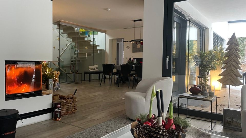 Offener, moderner Wohn- und Essbereich mit verglastem Kamin links, Holzfußboden und Treppe mit Glasgeländer im Hintergrund. Rechts führen große bodentiefe Schiebetüren auf eine Terrasse mit Pflanzen und dekorativer Beleuchtung.
