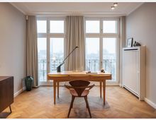 Helles Arbeitszimmer mit einem hölzernen Schreibtisch und Stuhl auf einem Fischgrätenparkettboden. Große Fenster mit Vorhängen bieten einen Blick auf eine städtische Umgebung mit Gebäuden, während dekorative Elemente wie ein Keramikgefäß und minimalistische Dekorationen den Raum vervollständigen.