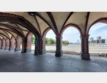 Gewölbter Durchgang mit gotischen Backsteinbögen, die auf steinernen Pfeilern ruhen. Im Hintergrund sind eine Straße, ein Fluss, moderne Gebäude und Bäume zu sehen.