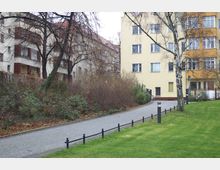 Gepflasterter Weg, der durch ein Wohngebiet mit mehrstöckigen beige- und hellgelben Gebäuden führt. Rechts im Bild befindet sich eine Rasenfläche, links Büsche und Bäume, teilweise kahl.