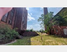 Industriegelände mit roten Backsteingebäuden, umgeben von Gras und vereinzelten Bäumen. Im Hintergrund ist ein hoher Schornstein sowie ein freier Himmel mit Wolken sichtbar.