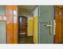 Ein schmaler Flur mit gelb-weiß gestrichenen Wänden, einer braunen Holztür mit schwarzen Beschlägen im Hintergrund und teilweise geöffneten Holztüren im Vordergrund, darunter eine graue Tür mit Metallklinke.