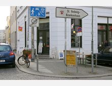 Straßenecke mit einem weißen Gebäude, darin Geschäfte mit großen Fenstern und einem dunklen Eingang. Schilder weisen auf das „St. Hedwig-Krankenhaus“ und eine 30er-Zone hin. Geparkte Autos, ein Fahrrad mit Korb, ein Werbeschild und ein Zeitungsständer befinden sich im Vordergrund.