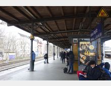 Überdachter Bahnsteig des S-Bahnhofs Rathaus Steglitz mit einer Holzdachkonstruktion und gusseisernen Stützpfeilern. Menschen warten entlang des Bahnsteigs nahe der Schienen, einige sitzen unter einem Werbeplakat, das an einer gelb gefliesten Wand hängt.