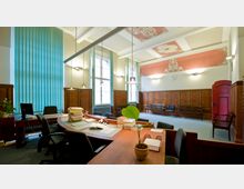 Geräumiges Büro mit hohen Decken, verzierten Holzvertäfelungen und großen Fenstern mit grünen Vertikaljalousien. Im Vordergrund stehen Schreibtische mit Aktenstapeln und Bürostühlen, im Hintergrund mehrere Wartebänke und eine rote Holztür.