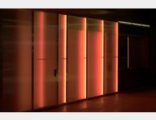 Ein Innenraum mit vertikalen, halbtransparenten Glaspaneelen, die in warmem orange-rotem Licht hinterleuchtet sind, vor einem dunklen Hintergrund.