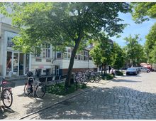 Straßenszene mit Kopfsteinpflaster, flankiert von kleinen Bäumen und geparkten Fahrrädern. Im Hintergrund befinden sich Geschäfte mit Schaufenstern, darunter ein Büro mit dem Schriftzug „ERGO“, sowie parkende Autos und ein Verkaufsstand.