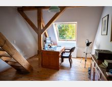 Dachgeschoss-Arbeitszimmer mit sichtbaren Holzbalken und Holzdielenboden, einem Schreibtisch mit Computer sowie einem Stuhl. Ein Dachfenster bringt Tageslicht herein; daneben steht ein Stativ, und an der rechten Wand befinden sich Möbel und gerahmte Bilder.