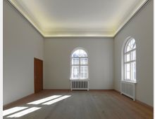 Ein leerer, heller Raum mit hohen Decken, großen bogenförmigen Fenstern und hellen Wänden. Der Raum verfügt über einen Holzfußboden, weiße Heizkörper unter den Fenstern und eine Holztür auf der linken Seite.