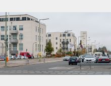 Straßenszene in einem städtischen Wohngebiet mit modernen mehrstöckigen Wohngebäuden im Hintergrund, großen Fenstern und Balkonen. Im Vordergrund stehen parkende und fahrende Autos auf einer breiten Straße, flankiert von Laternen und vereinzelten Bäumen.