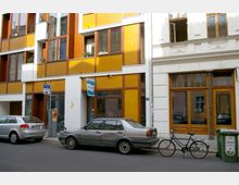 Straßenszene mit modernen Wohn- und Geschäftshäusern, einer Fassade mit großen gelben und weißen Flächen sowie braunen Fensterrahmen. Vor dem Gebäude parken ein silbernes Auto und ein Fahrrad, daneben steht eine grüne Glasrecycling-Tonne.