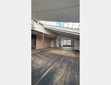 Großer, leerer Loft- oder Lagerraum mit abgenutztem Holzboden, Sichtziegelwänden und schräger Decke mit Oberlichtfenstern. An der Rückwand stehen mehrere rechteckige Fenster über einem Heizkörper, daneben eine Tür und ein Feuerlöscher.