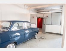 Innenraum einer hellen Werkstatt- oder Garagenhalle mit weiß gestrichenen Wänden, sichtbaren Deckenrohren und abgenutztem Betonboden. Links steht ein dunkelblauer Oldtimer, rechts sind verglaste Trennwände mit einer offenen Tür zu einem roten Nebenraum zu sehen.