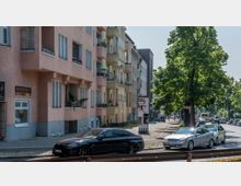 Straßenszene mit mehrstöckigen Wohnhäusern im Vordergrund, die pastellfarbene Fassaden und Balkone haben, auf denen Pflanzen stehen. Rechts der Straße stehen geparkte Autos unter großen grünen Bäumen, während im Hintergrund weitere Gebäude und fahrende Autos zu sehen sind.